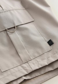 Tissu beige avec une poche rectangulaire comportant un rabat, des détails cousus, un petit anneau métallique et une étiquette logo triangulaire noire.