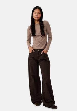 Vrouw staat met handen in de zakken, draagt een taupekleurige top met lange mouwen en een wijde donkerbruine broek tegen een effen achtergrond.