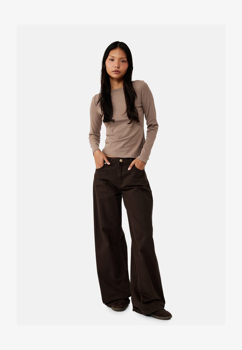 Mujer de pie con las manos en los bolsillos, vistiendo una blusa de manga larga color topo y pantalones anchos de color marrón oscuro sobre un fondo liso.