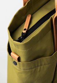 Olive green tote bag with tan leather accents, zippered pocket, and stitched details. Features a smooth texture and multiple external pockets.