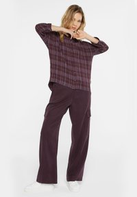Oversized burgundy plaid button-up shirt with long sleeves and white buttons, paired with loose dark pants featuring cargo pockets and white sneakers.