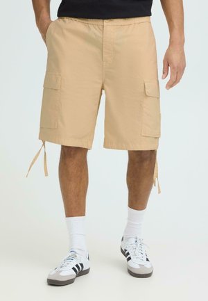 Mann iført beige cargoshorts med store sidelommer, hvite crew-sokker og hvite joggesko med svarte striper, stående mot en hvit bakgrunn.