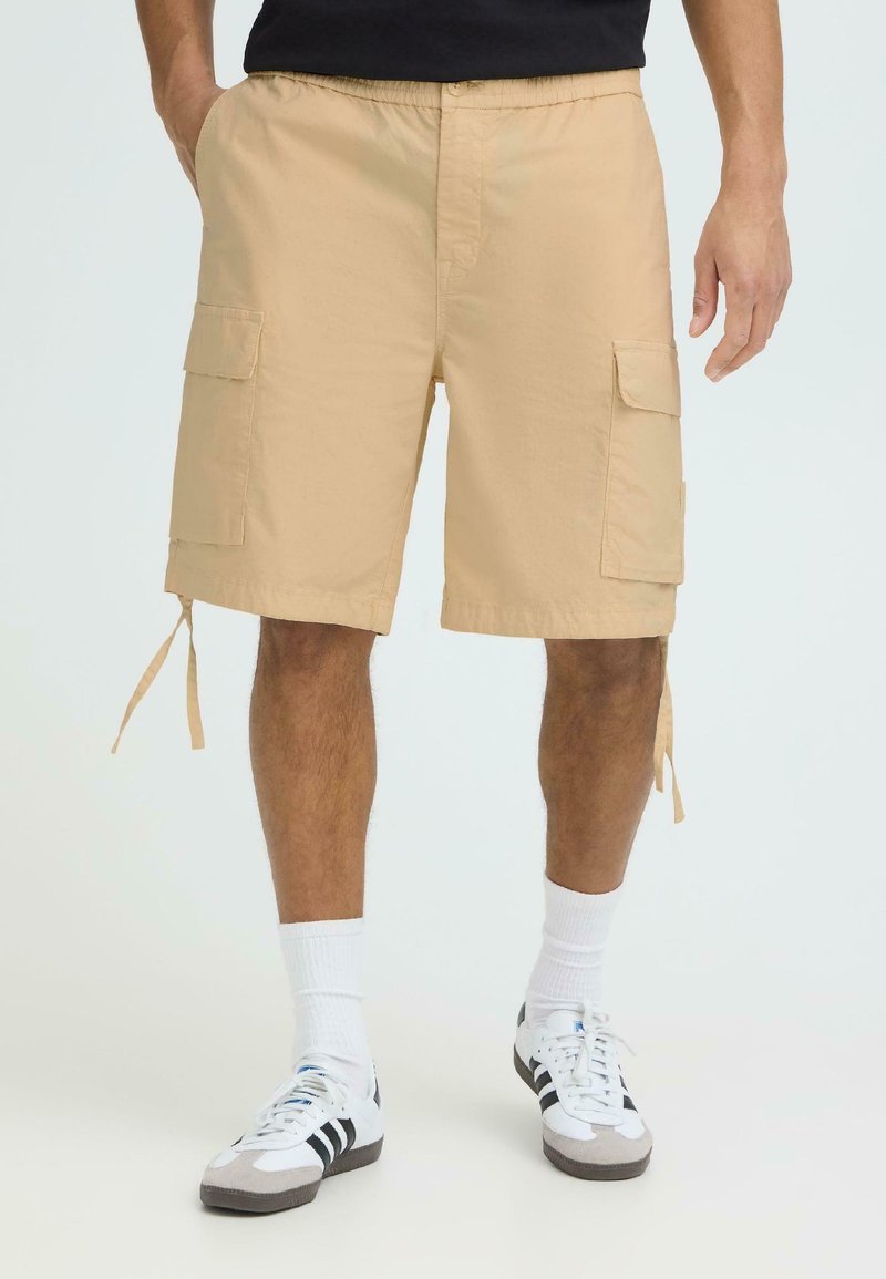 Homme portant un short cargo beige avec de grandes poches latérales, des chaussettes blanches montantes et des baskets blanches à rayures noires, debout devant un fond blanc.