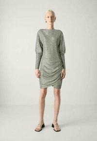 Robe métallisée argentée avec des manches longues, des épaules bouffantes et un devant drapé. Texture lisse et design jusqu'aux genoux. Portée avec des sandales plates.