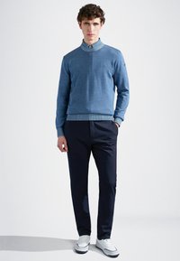 Maglione azzurro chiaro a maglia con colletto alto, abbinato a pantaloni blu scuro. Il maglione presenta polsini a coste e una texture liscia. Sneakers bianche completano l'outfit.