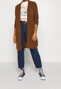 Brązowy sweter z dzianiny z otwartym przodem, długimi rękawami, bocznymi kieszeniami i luźnym krojem, zestawiony z koszulką z nadrukiem, niebieskimi dżinsami i czarnymi trampkami.