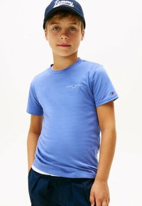 Ragazzo che indossa una maglietta blu di Tommy Hilfiger e un berretto navy, in piedi con le mani nelle tasche di pantaloni scuri contro uno sfondo bianco semplice.