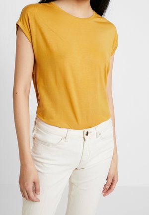 Kobieta w musztardowożółtym krótkim rękawem wsuniętym w wysokowatowe ecru jeansy z przodu z guzikami i kieszeniami.