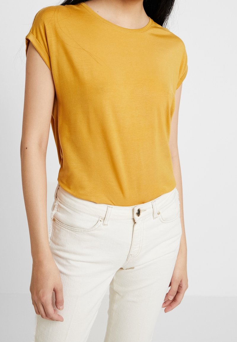 Femme portant un haut jaune moutarde à manches courtes tucked dans un jean taille haute blanc cassé avec bouton frontal et poches.