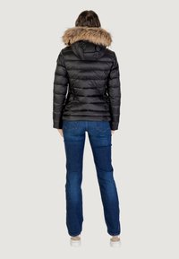 Svart puffjacka med huva i konstpäls, quiltad design och figurnära silhuett, kombinerad med blå denimjeans och ljusa skor.