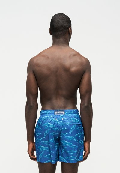 Άνδρας με σκούρο χρώμα δέρματος, φαίνεται από πίσω, φοράει μπλε μαγιό με σχέδιο φάλαινας πάνω σε απλό ανοιχτό φόντο.