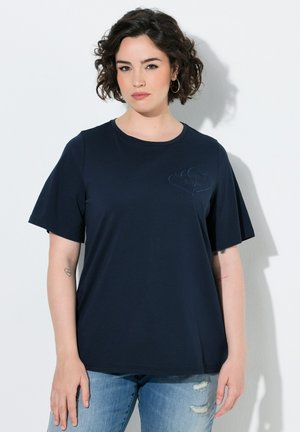 Ung kvinde med kort krøllet hår iført en navy t-shirt med hjertedesign og revnede blå jeans, står mod en hvid væg.