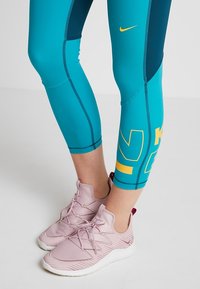 Ben i petrolblå och marinblå beskurna träningsleggings med gula detaljer, iförda ljusrosa snörskor med vita sulor på ett rent golv.