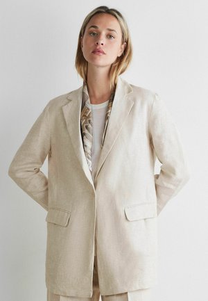 Kobieta w beżowej lnianej marynarce na białej bluzce z wzorzystym szalikiem schowanym do środka, stojąca na jednolitym białym tle.