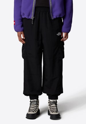 Osoba ubrana w czarne spodnie typu cargo z logo The North Face, beżowe buty trekkingowe i fioletową bluzę polarową, stojąca na jasnym tle.