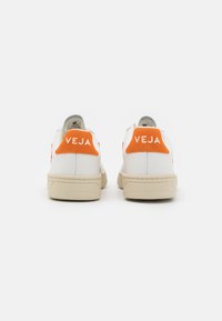 Baskets en cuir blanc avec des accents de talon en suede orange, dotées d'une semelle plate crème et de coutures visibles, avec un logo à l'arrière.