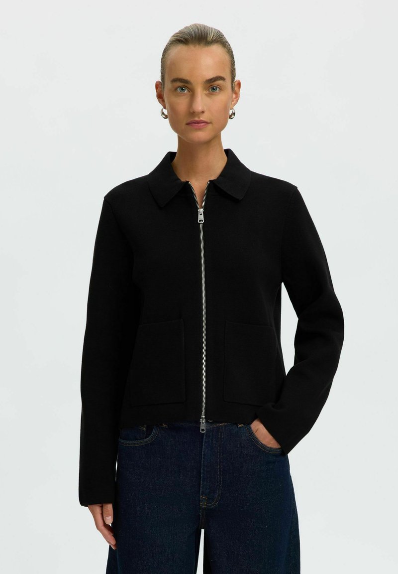 Femme aux cheveux tirés en arrière portant une veste noire zippée avec col et grandes poches à l’avant sur un jean bleu foncé, debout devant un fond uni.