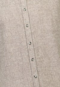 Camisa de lino color marrón claro con un frente abotonado con botones plateados, tejido texturizado y detalles de costura vertical.