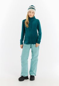 Jong persoon in een teal capuchonjack, lichtblauwe broek, zwarte laarzen en een witte en teal beanie met tekst, staand op een witte achtergrond.