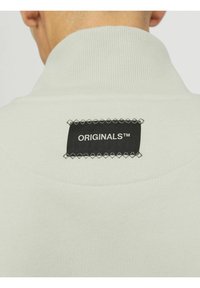 Lys grå sweatshirt med høj krave, prydet med en sort rektangulær label, der står "ORIGINALS™," omkranset af dekorativ syning.