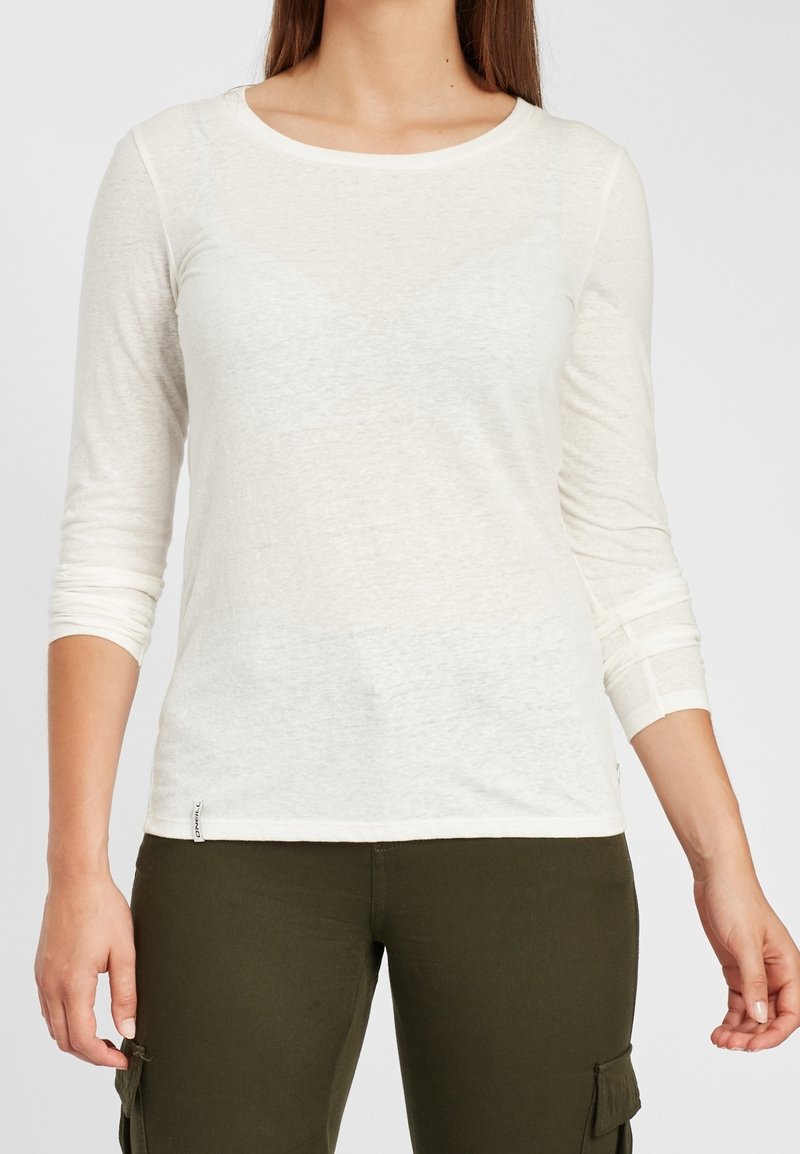 Leichtes, langärmliges Oberteil in Off-White mit einem Rundhalsausschnitt, strukturiertem Stoff und einem kleinen Etikett am Saum, kombiniert mit olivgrünen Hosen.