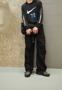 Personne portant un pantalon cargo noir, des baskets noires et grises, et un t-shirt à manches longues noir avec "72" et le swoosh de Nike, tenant un sac noir.