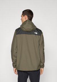 Waterdichte jas in olijfgroen met een zwart bovenpaneel, capuchon, elastische manchet en een logo op de achterkant. Gladde textuur, casual ontwerp.