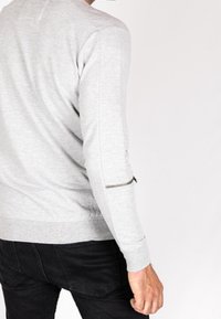 Sudadera gris de manga larga, con detalle de cremallera en el brazo izquierdo y dobladillo acanalado. Combinada con jeans oscuros.