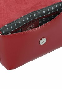 Bolso de cuero rojo con un acabado liso, que cuenta con un cierre a presión plateado y un forro de tela estampada que muestra la letra 'P'.