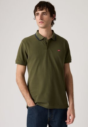 Młody mężczyzna ubrany w oliwkowo zieloną koszulkę polo z małym czerwonym logo Levi's oraz ciemnoniebieskie jeansy, stojący z jedną ręką w kieszeni.