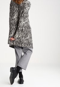 Randig jacka i stor storlek, svart och vit; grå byxor; svarta skor; textil med struktur och långa ärmar; avslappnad passform med sidofickor.