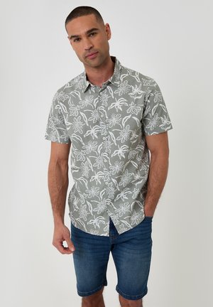 Hombre con camisa gris de manga corta con botones y estampados florales blancos, con pantalones cortos vaqueros azul oscuro, de pie frente a un fondo liso.