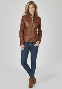 Braune Lederjacke mit einem Reißverschluss auf der Vorderseite, zwei Brusttaschen und einem Stehkragen. Kombiniert mit dunklen Jeans und braunen Knöchelstiefeln.