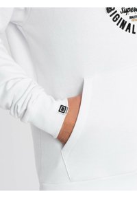 Sudadera blanca con un bolsillo delantero y puños acanalados. Presenta una pequeña etiqueta de logo en el puño y un diseño circular en el pecho.