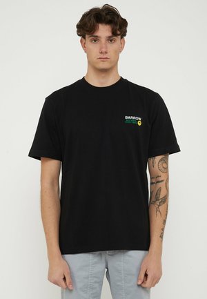 Giovane uomo con capelli corti e ricci, indossa una maglietta nera con la scritta "BARROW BAD VIBES GOOD TIMES" e pantaloni grigio chiaro, braccio sinistro tatuato visibile.