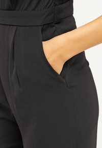Main placée dans la poche latérale d'un pantalon noir taille haute avec des plis près de la ceinture.