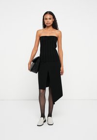 Top negru fără bretele, cu detaliu legat, asociat cu o fustă asimetrică neagră. Ciorapi negri și pantofi ușor gri, tip pantof de interior.