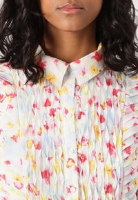Κοντομάνικο πουκάμισο με λουλουδάτο μοτίβο που χαρακτηρίζεται από μαλακή, πλισέ υφή, ελαφρύ ύφασμα, κλείσιμο με κουμπιά και παστέλ παλέτα χρωμάτων με κόκκινες, κίτρινες και μπλε αποχρώσεις.