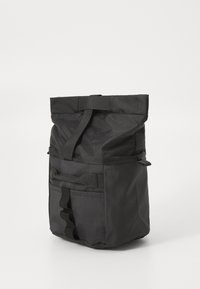 Sac à dos noir à rabat avec poche avant, sangle à boucle et boucles latérales sur fond blanc.