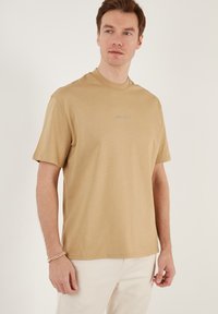Beige bomulds t-shirt med rund hals og korte ærmer. Har et diskret blåt broderet logo på brystet. Glat tekstur.