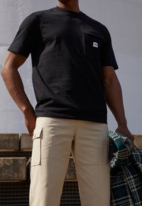 T-shirt noir à manches courtes avec une poche frontale et un patch logo, associé à un pantalon cargo beige avec des poches latérales, tenant une veste à carreaux verte.
