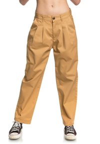 Khaki plisované kalhoty vyrobené z lehkého materiálu s volným střihem, vybavené knoflíkovým zapínáním a bočními kapsami, spárované s černými teniskami.