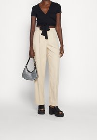 Černý zavinovací top s krátkými rukávy, béžové kalhoty s vysokým pasem, robustní černé sandály na platformě a malá kabelka se stříbrnými akcenty.