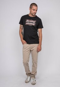 T-shirt en coton noir avec un graphisme "BLUE & JEANS", associé à un chino beige clair et des baskets blanches. Design simple et coupe décontractée.