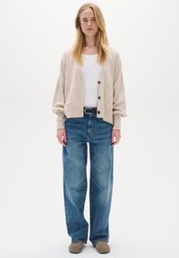 Žena s dlhými blond vlasmi, oblečená v béžovom kardigane, bielom tričku, širokých modrých džínsoch, čiernom opasku a hnedých topánkach stojaca na bielom pozadí.