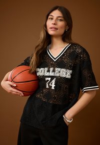 Mlada ženska drži košarkarsko žogo v roki, oblečena v črno čipkasto majico z napisom "COLLEGE 74" in črne hlače, na rjavi podlagi.