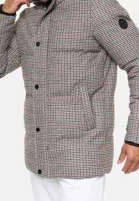 Toppatakki harmaassa, mustassa ja punaisessa ruutukuosissa; edessä vetoketju, korkea kaulus ja mustat yksityiskohdat hihoissa ja taskussa.