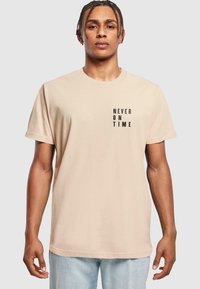 Beige bomulds-t-shirt med korte ærmer, der har teksten "ALDRIG TIL TID" i sort, placeret på venstre side.