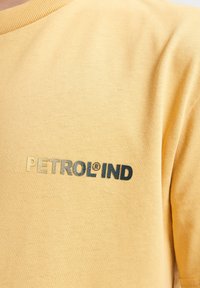 Gul bomulds T-shirt med ribbet rund hals og et trykt logo i guld og mørkeblå. Teksten "PETROL®IND" fremhæves.