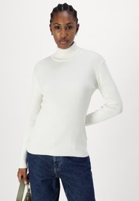 Pull col roulé blanc à côtes, à manches longues, au design ajusté, au toucher doux, porté avec un jean bleu foncé et tenant un sac à main vert.